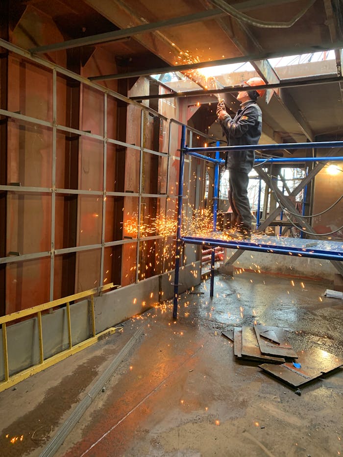 A construction worker welds metal indoors, creating flying sparks in a factory setting.