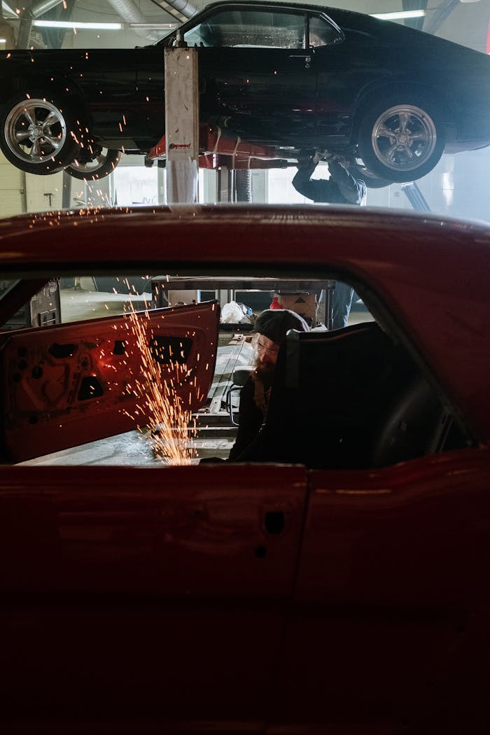 A mechanic welding inside a car workshop with a vehicle on a lift above.